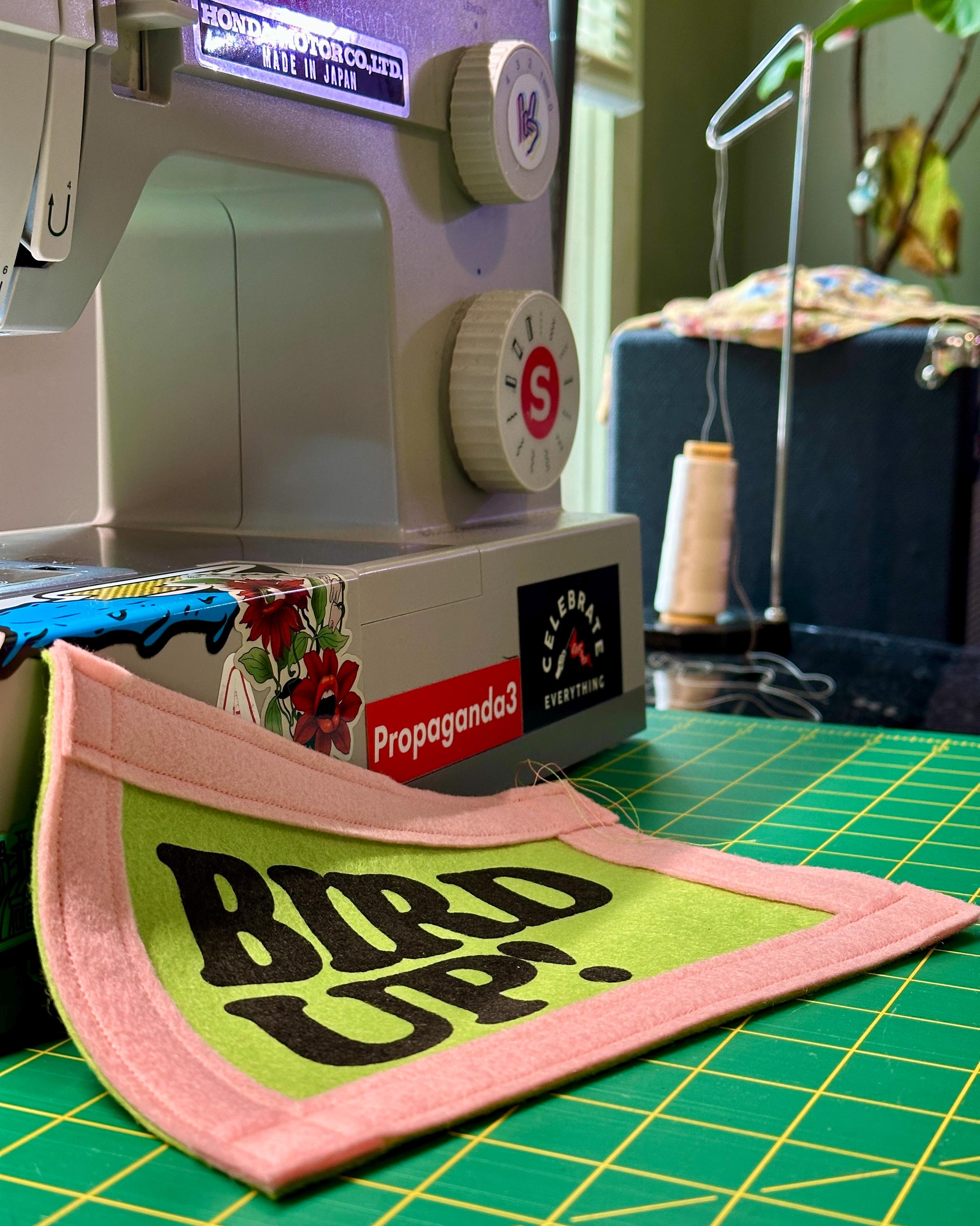 Sewing machine with a green and pink felt banner displaying 'Bird Up!' on a green cutting mat.