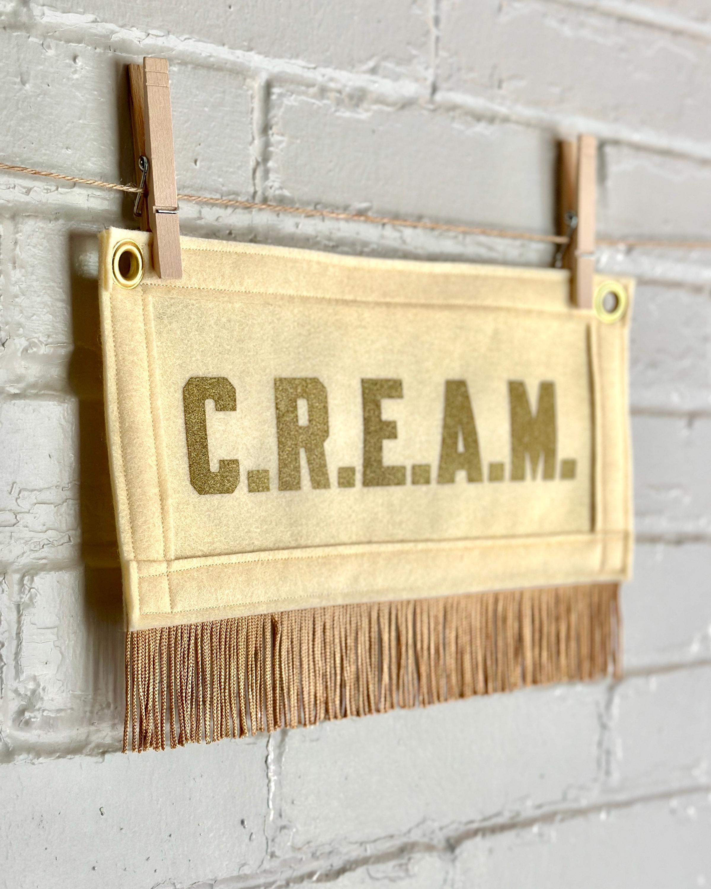 Side view of a cream felt banner with cream trim and gold fringe, and gold lettering that reads 'C.R.E.A.M.'