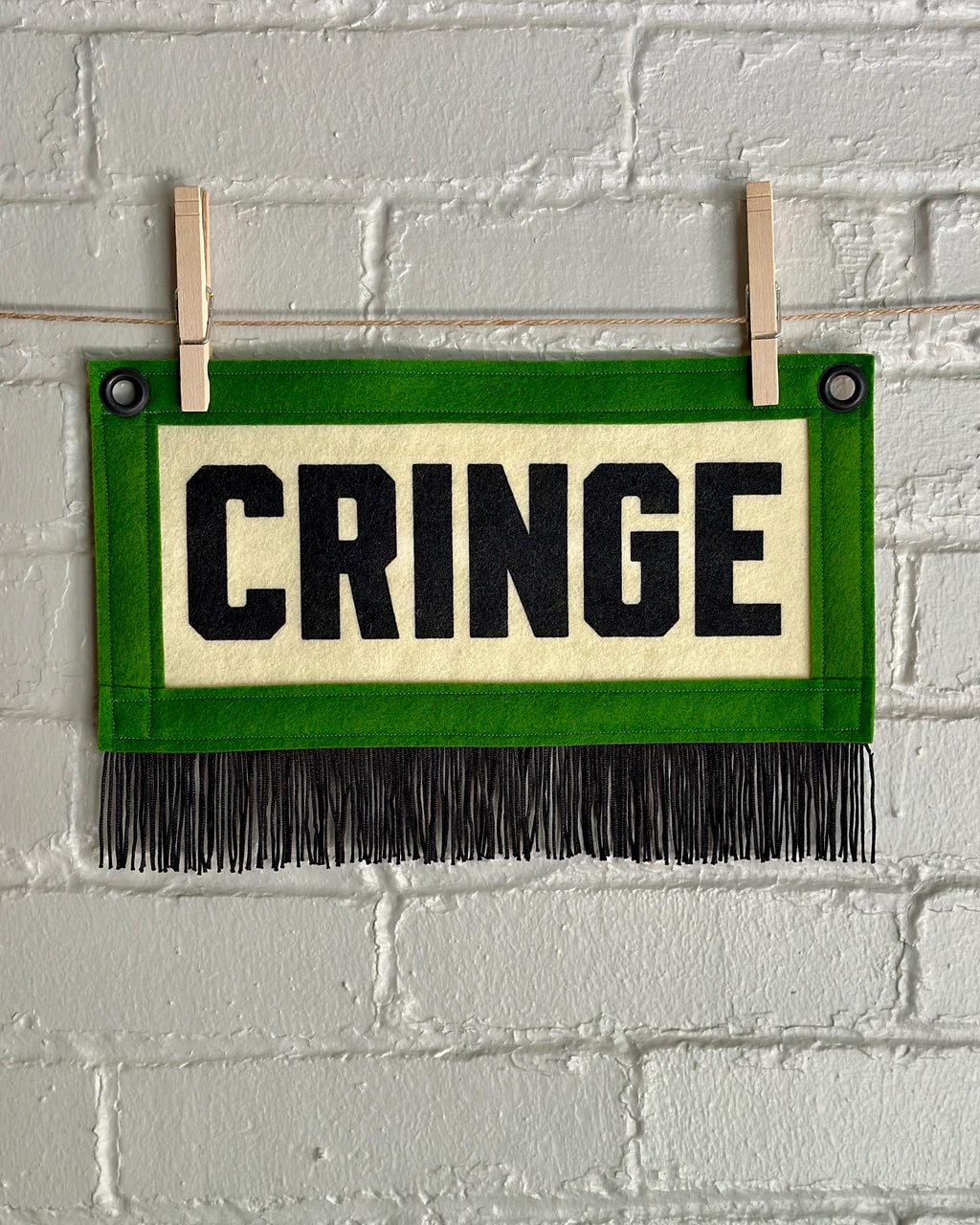 A cream felt banner with green trim, black fringe, and black lettering that reads 'CRINGE'