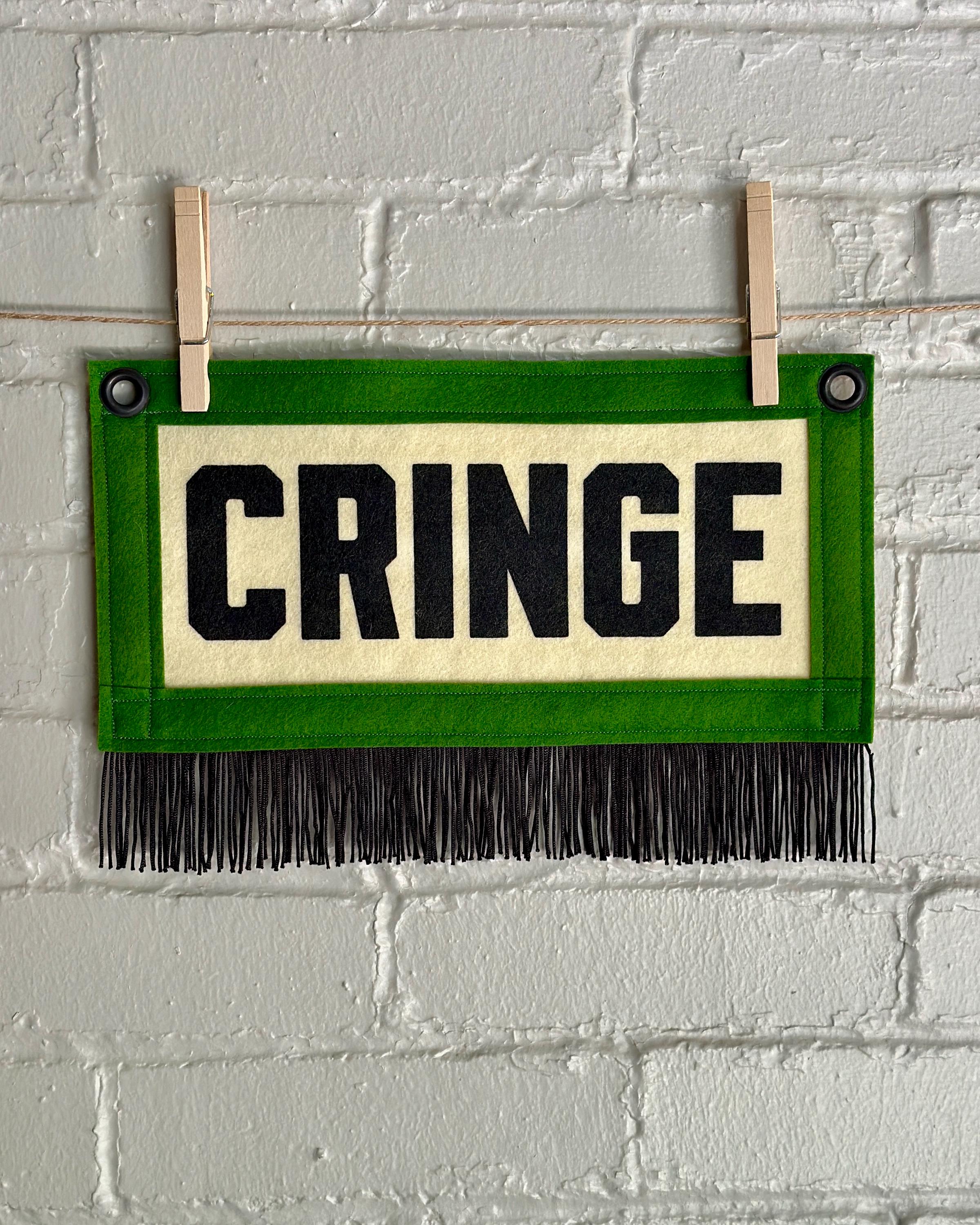A cream felt banner with green trim, black fringe, and black lettering that reads 'CRINGE'