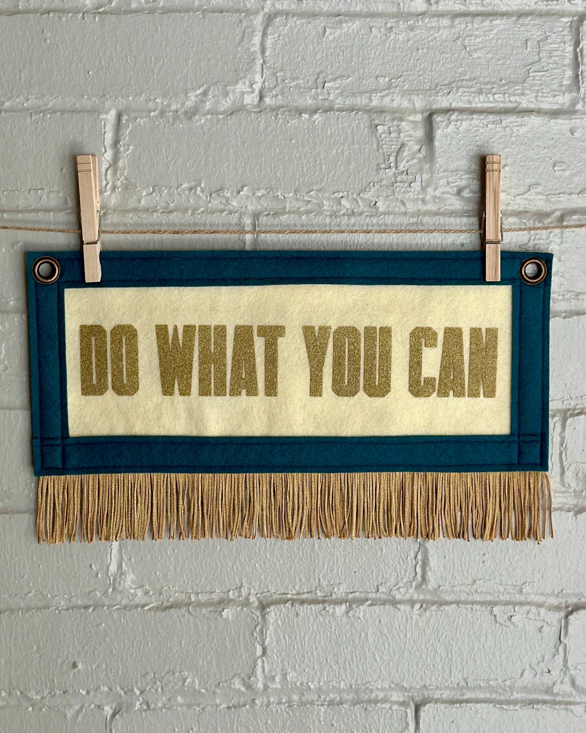A cream felt banner with blue spruce trim, gold fringe and lettering that reads "Do what you can"