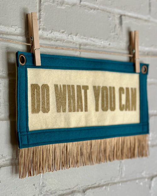 Side view of a cream felt banner with blue spruce trim, gold fringe and lettering that reads "Do what you can"