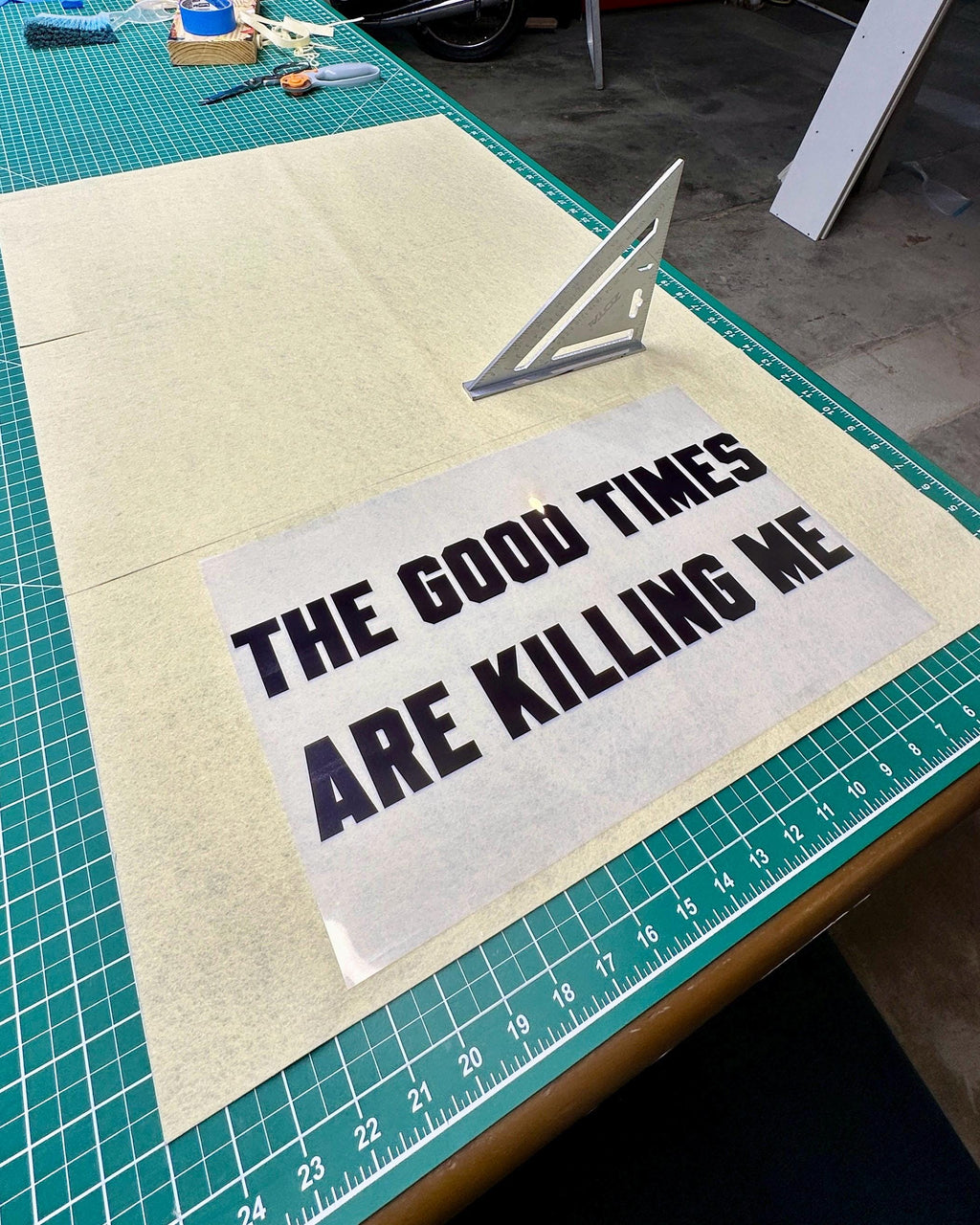 Three slices of cream felt on a large cutting table with a carpenter square and a transparency sheet with "the good times are killing me" printed on