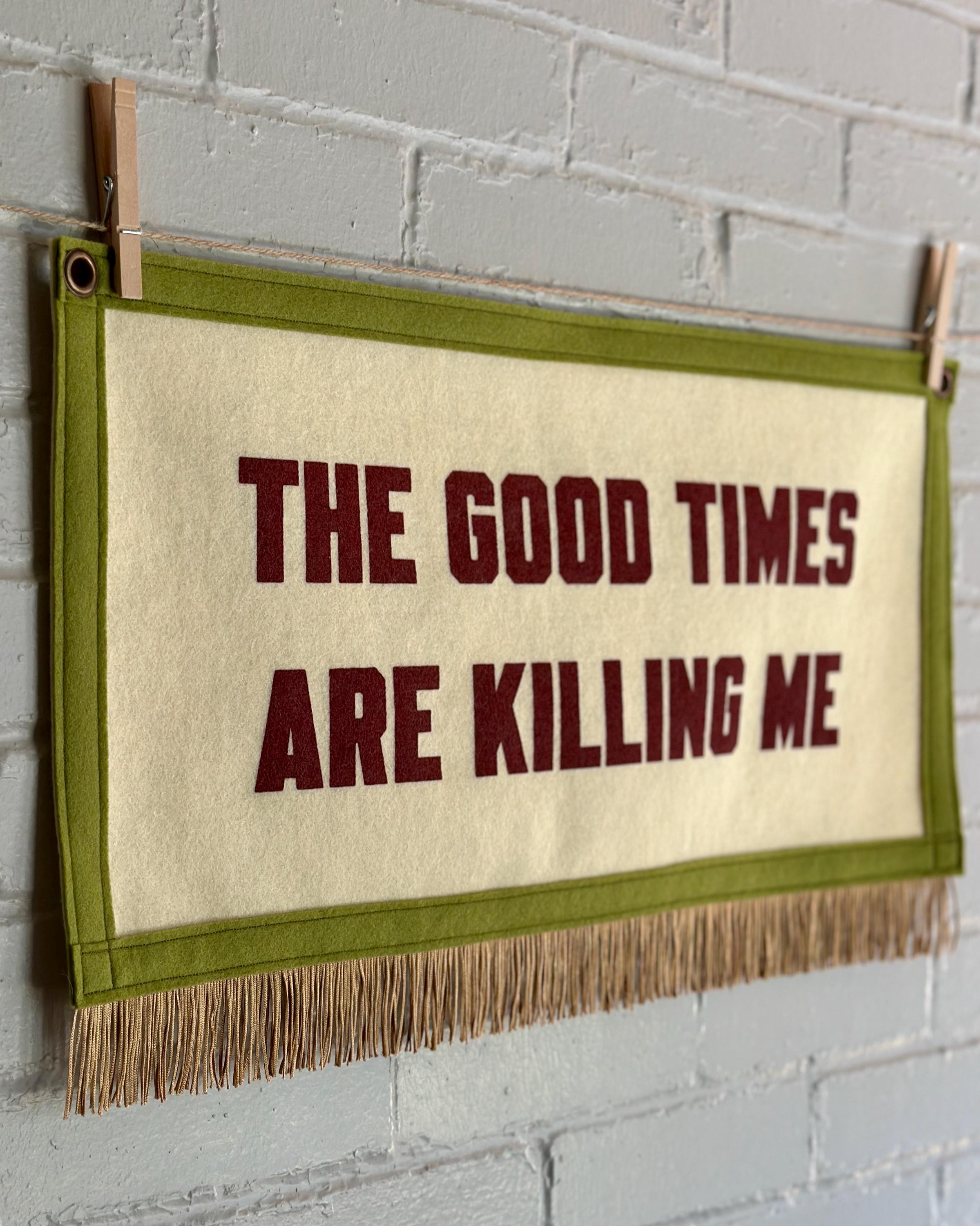Side view of a large cream banner with pea green felt trim, gold fringe, and blood red lettering that reads "The good times are killing me"