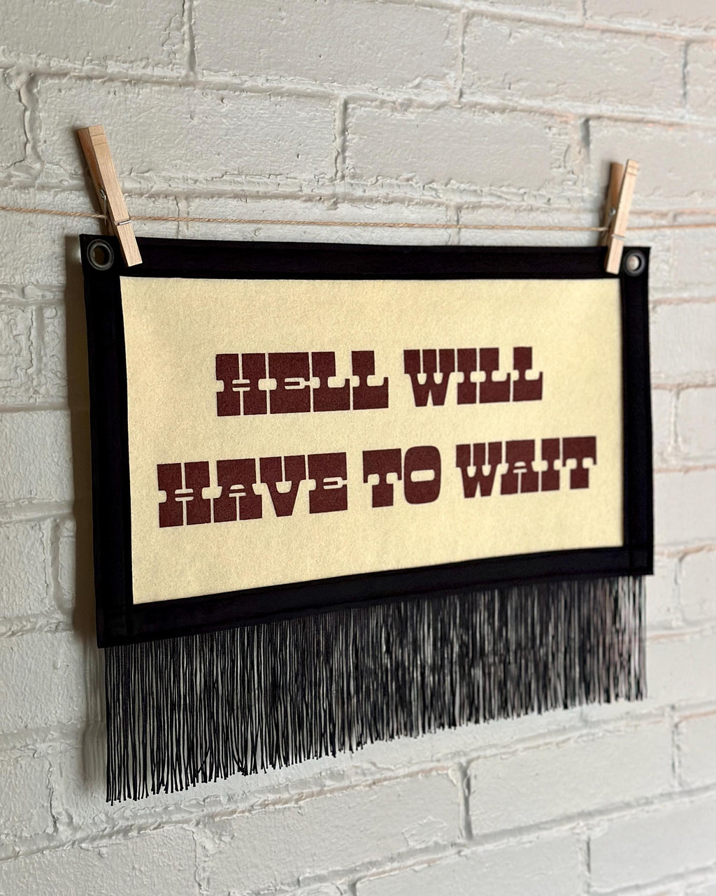 Side view of a cream felt banner with black trim, long black fringe, and blood red lettering reading "Hell will have to wait"