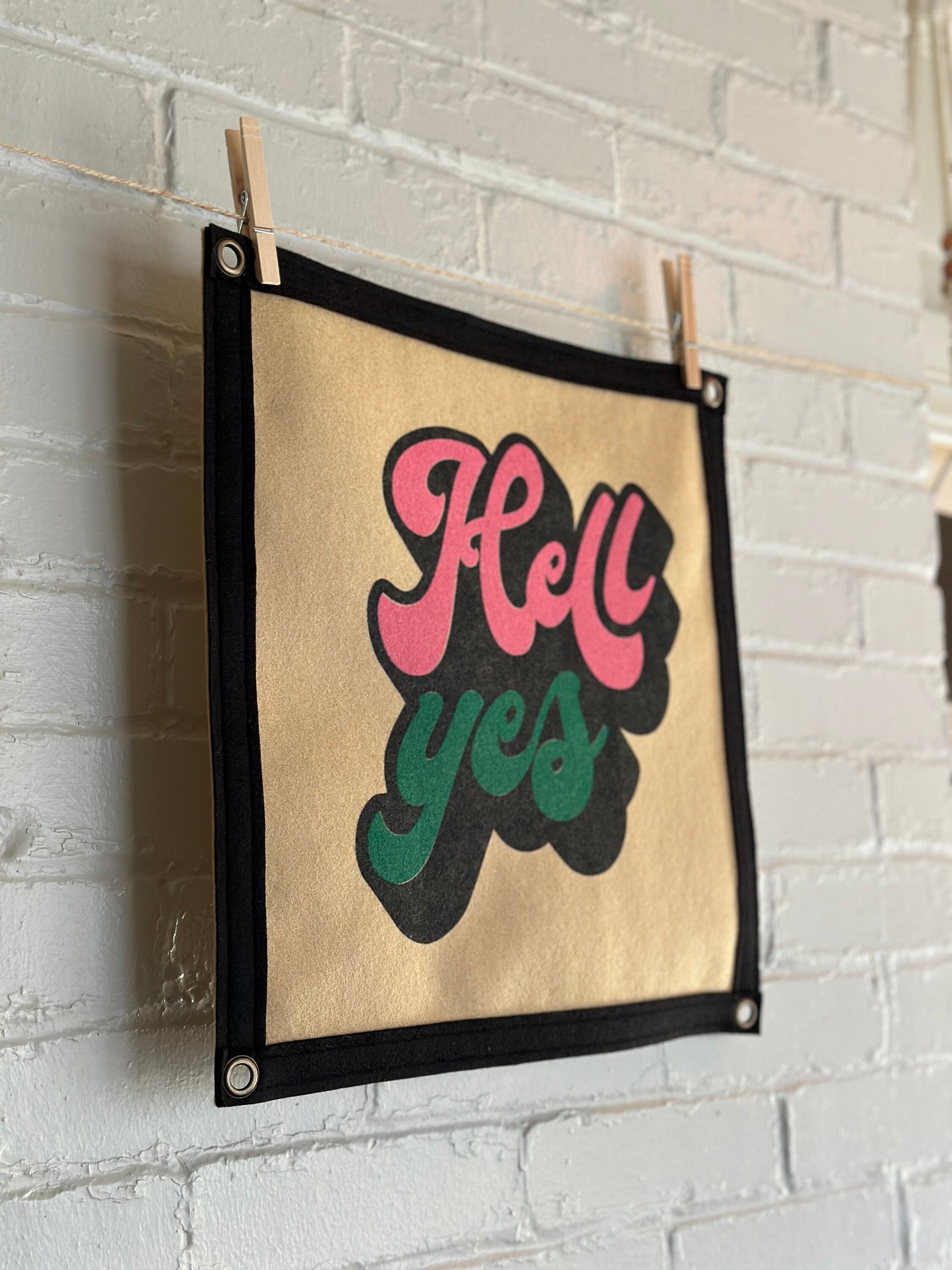 Side view of a light brown banner with black trim and a large stylized vintage lettering in black, pink and green ink that reads "Hell yes"
