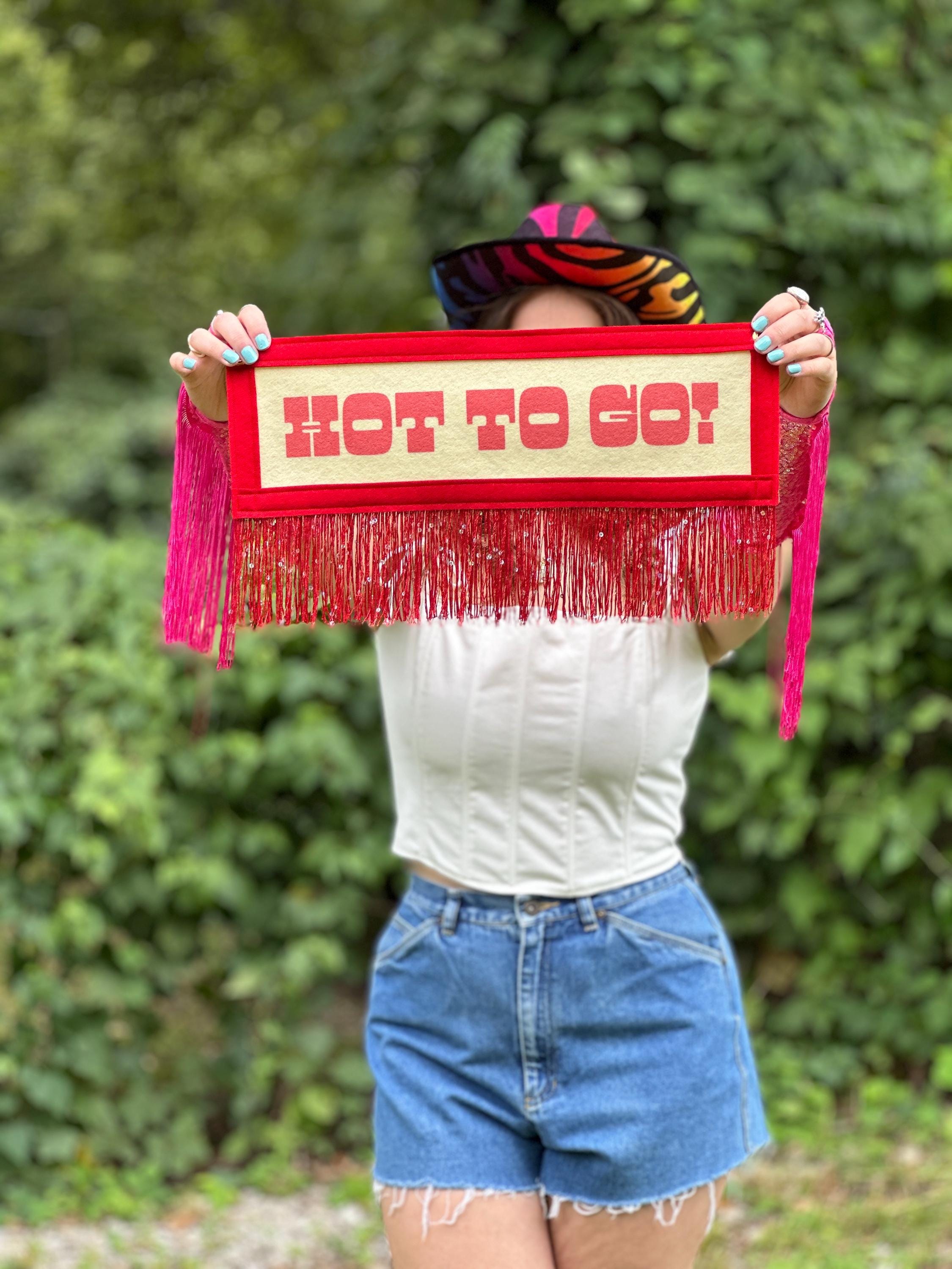 A woman with pink fringey gloves holding a red HOT TO GO! banner near the camera in an outdoor setting