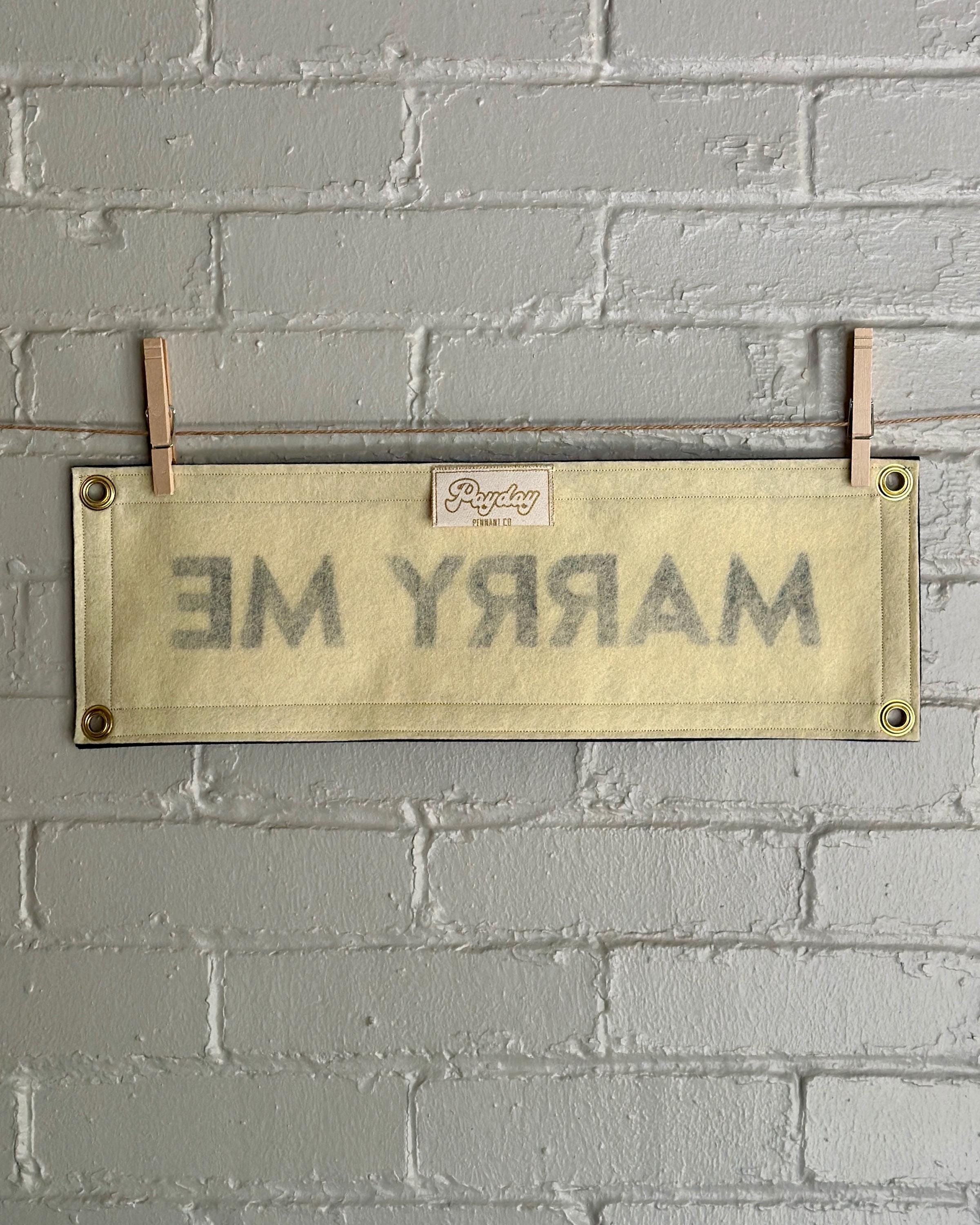 Back of a cream felt banner with black trim and lettering reading 'MARRY ME'