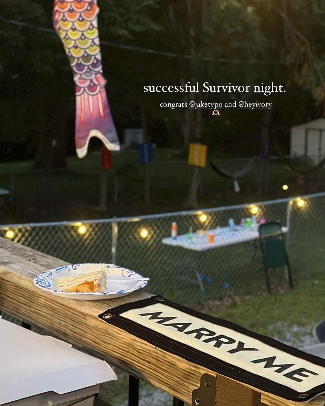 Table with 'Marry Me' sign, food, and 'Successful Survivor Night' text in an outdoor setting.