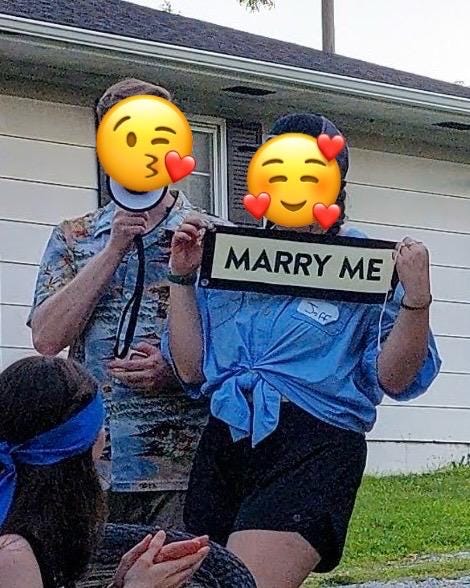 Two people holding a 'MARRY ME' sign in front of a house.