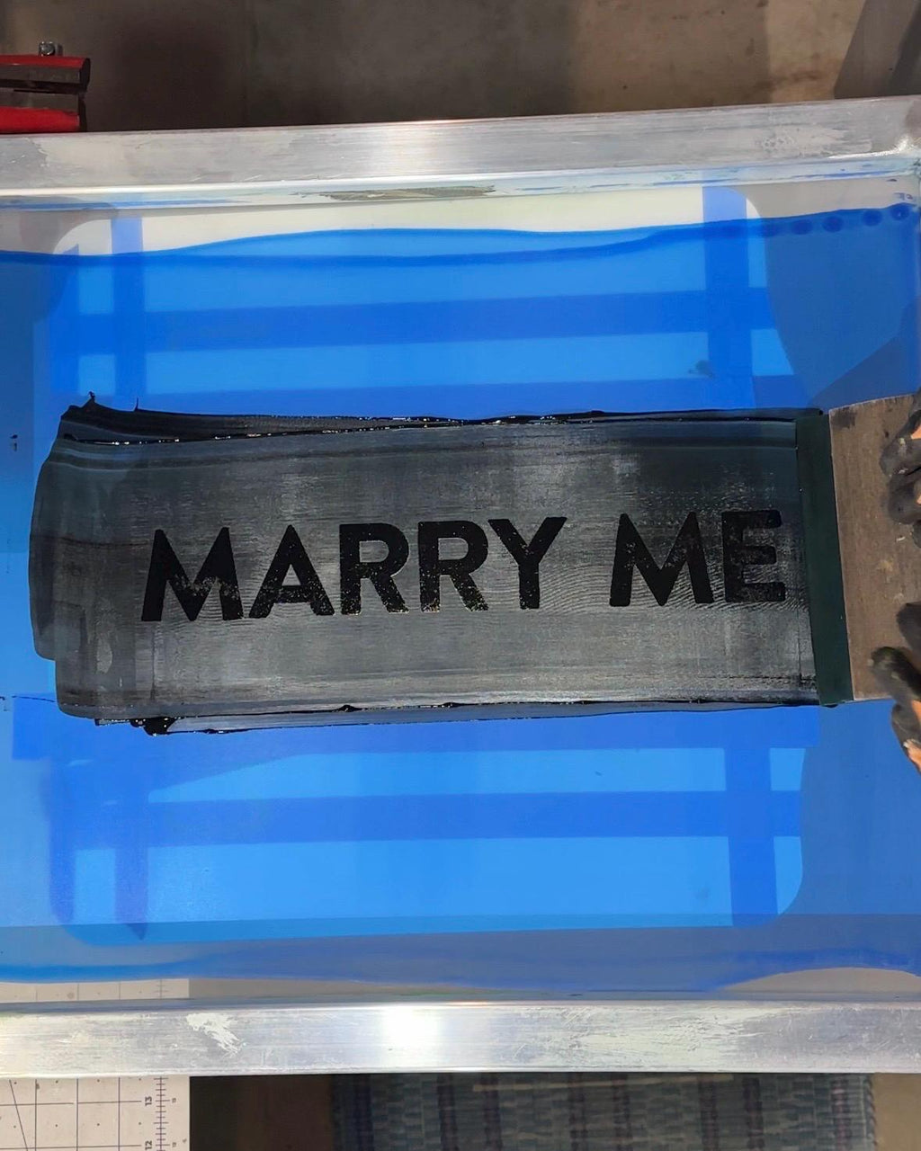 Silkscreen with 'Marry me' design and black ink being pulled by a squeegee on it