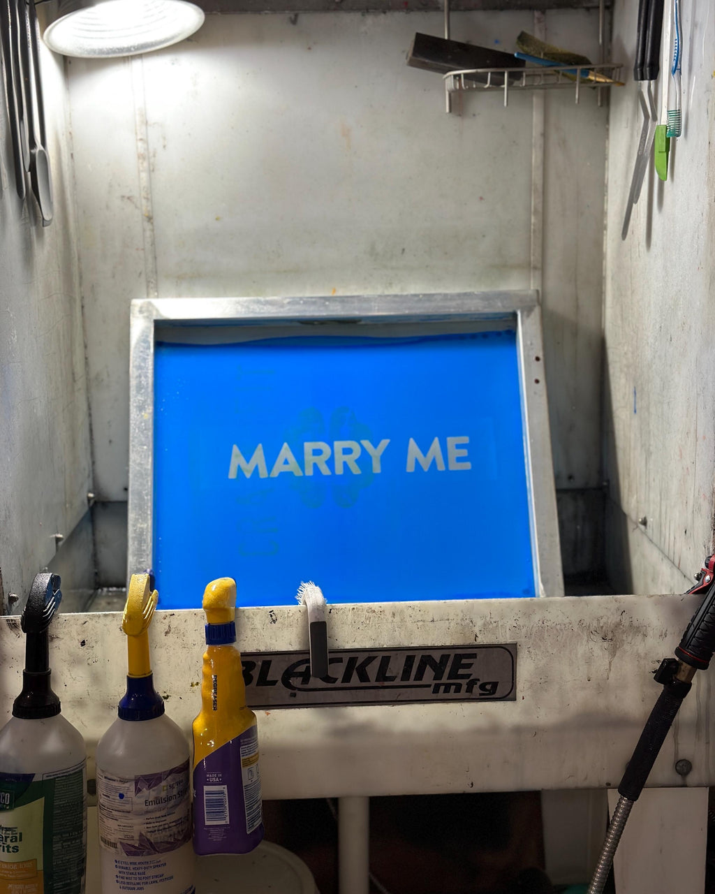 A blue silkscreen in a washout booth showing 'MARRY ME' text in a workshop setting