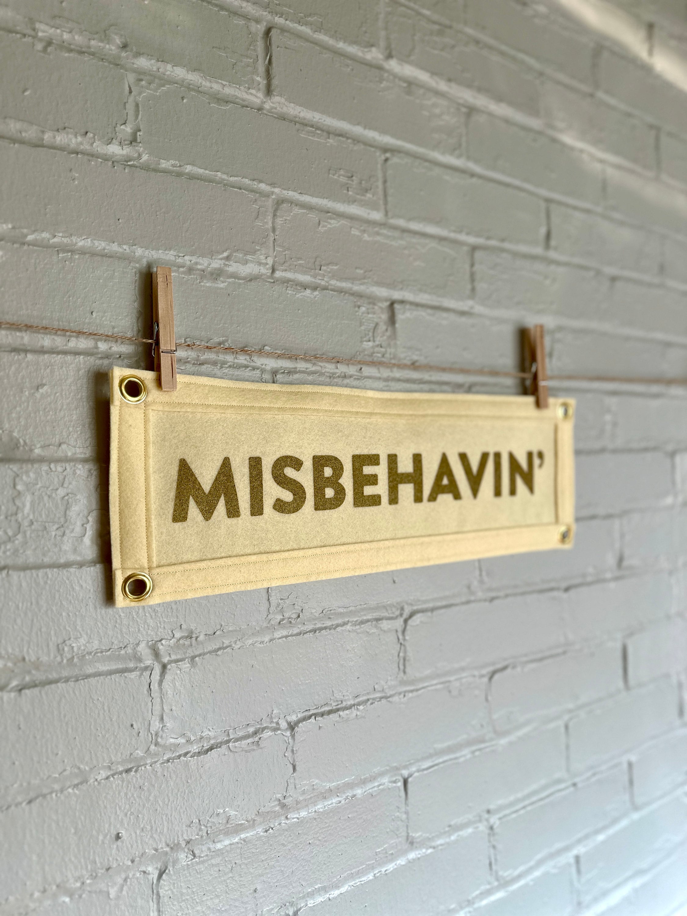 Side view of a cream felt banner with cream trim and gold ink lettering that reads "MISBEHAVIN'"