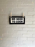 A cream felt banner with black trim and lettering that reads "we need to cook"