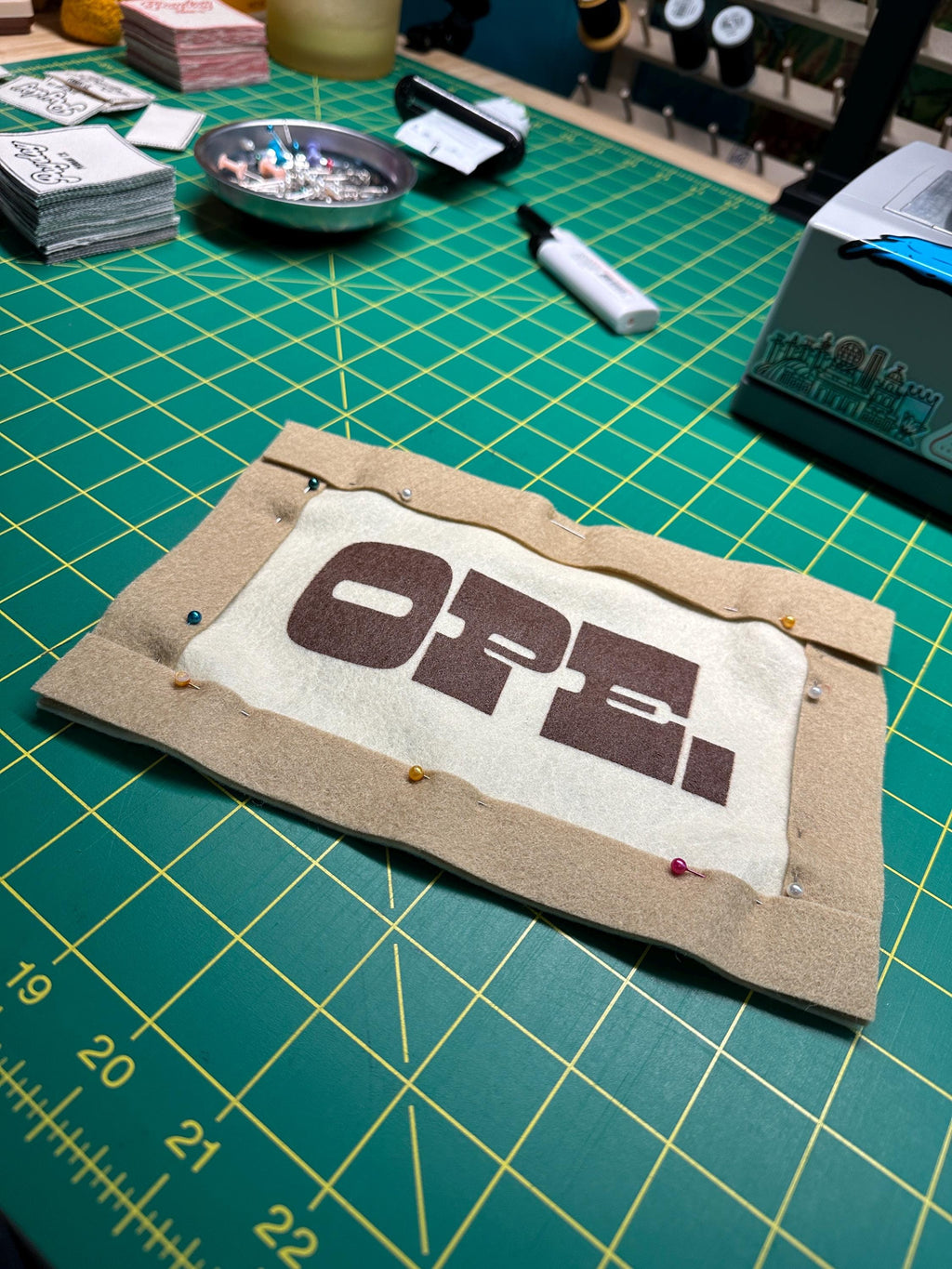 A felt banner in progress on a busy desk, printed with the word OPE and with brown trim pinned to the edges