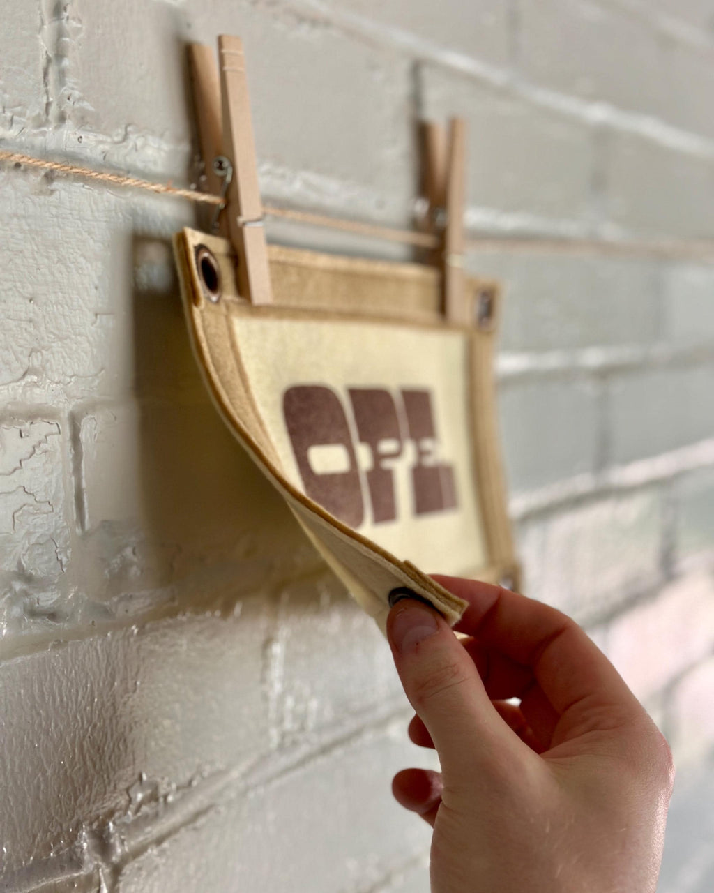 Side view with a hand pinching the corner of a cream felt banner with light brown trim and brown ink reading "OPE."