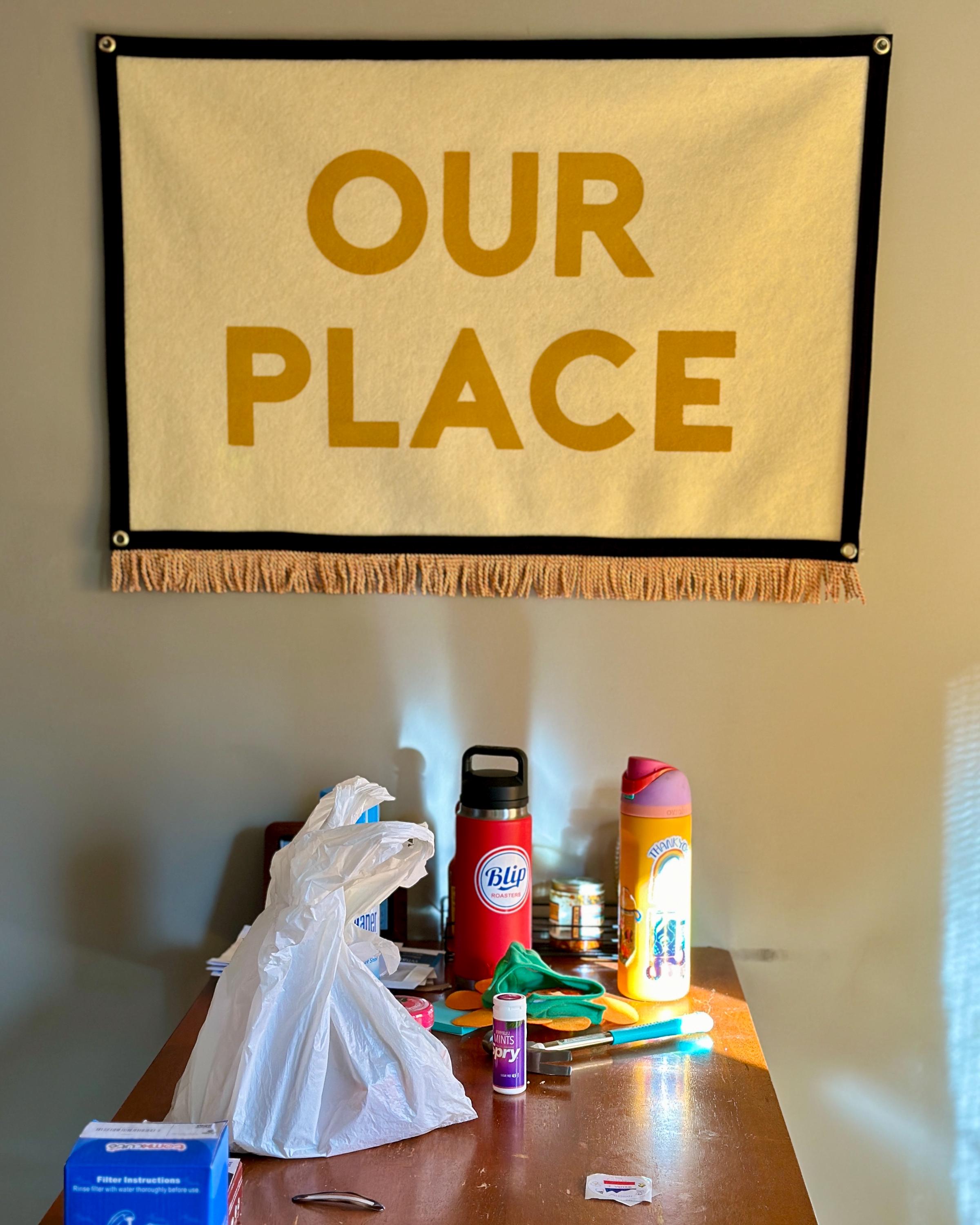 A dining room wall with cluttered table and above, a large cream felt banner with black trim, gold bullion fringe, and letters in yellow ink that read "OUR PLACE"