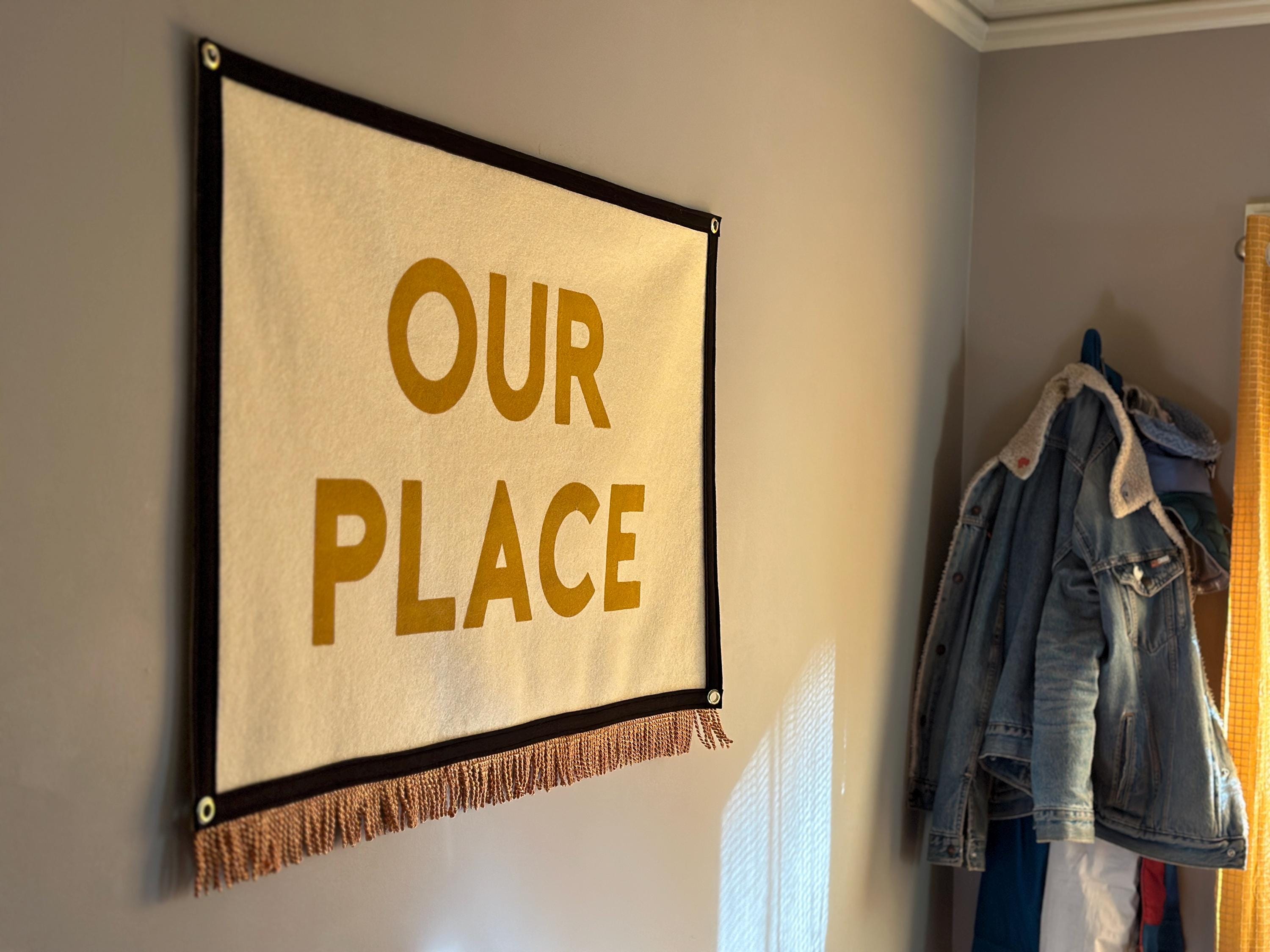 A dining room wall with coat rack and large cream felt banner with black trim, gold bullion fringe, and letters in yellow ink that read "OUR PLACE"