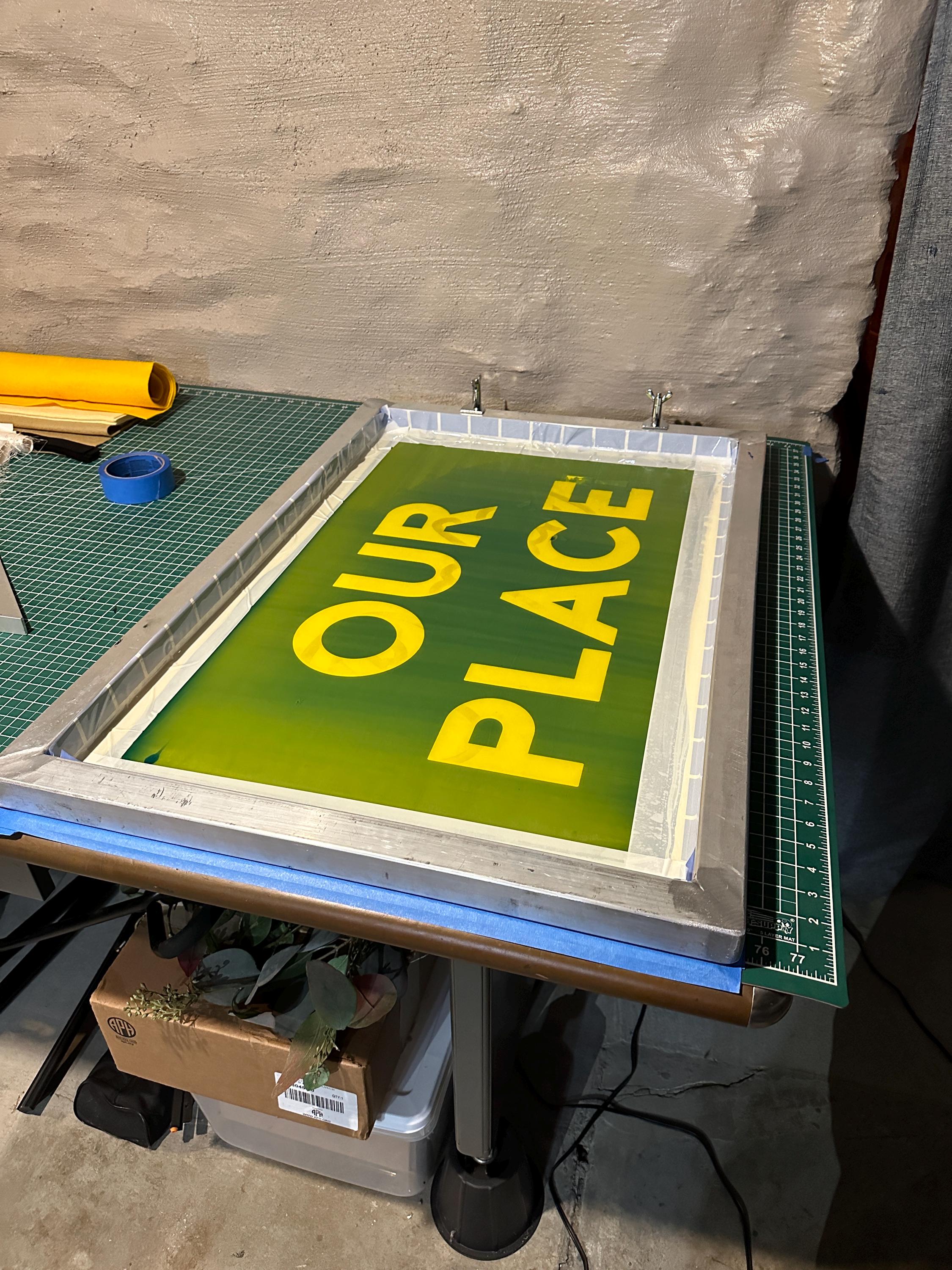A large silkscreen clamped to a table with the words "OUR PLACE" burned in and ready to be printed with