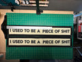 Two cream felt banners with black trim lettering reading "I USED TO BE A PIECE OF SHIT" sitting on top of a cutting mat. A T square is depicted showing a slightly crooked misaligned print