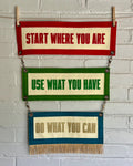 Three colorful felt banners with motivational phrases attached by brass rings, hanging on a white brick wall