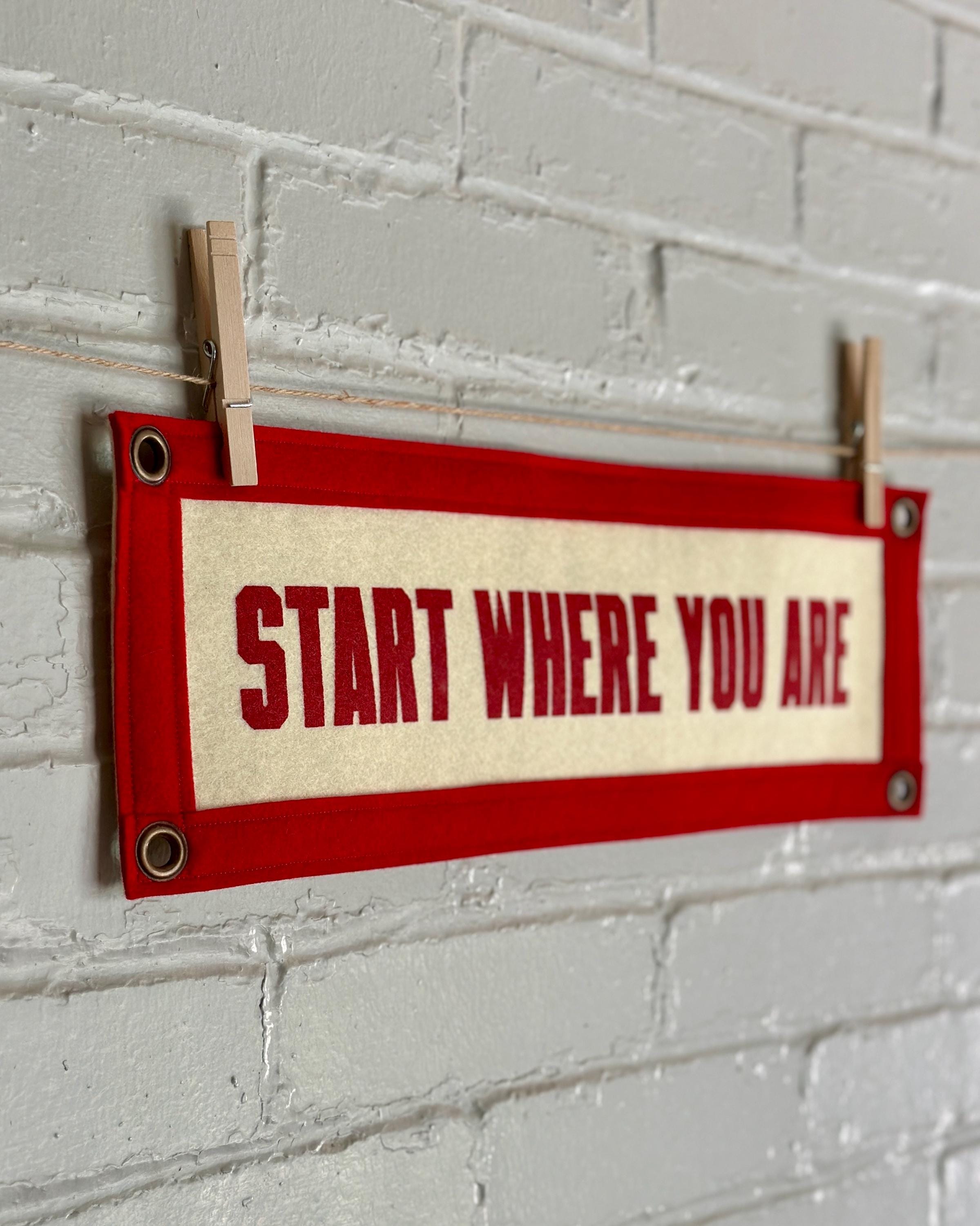 Side of a cream felt banner with red trim and lettering that reads "Start where you are"