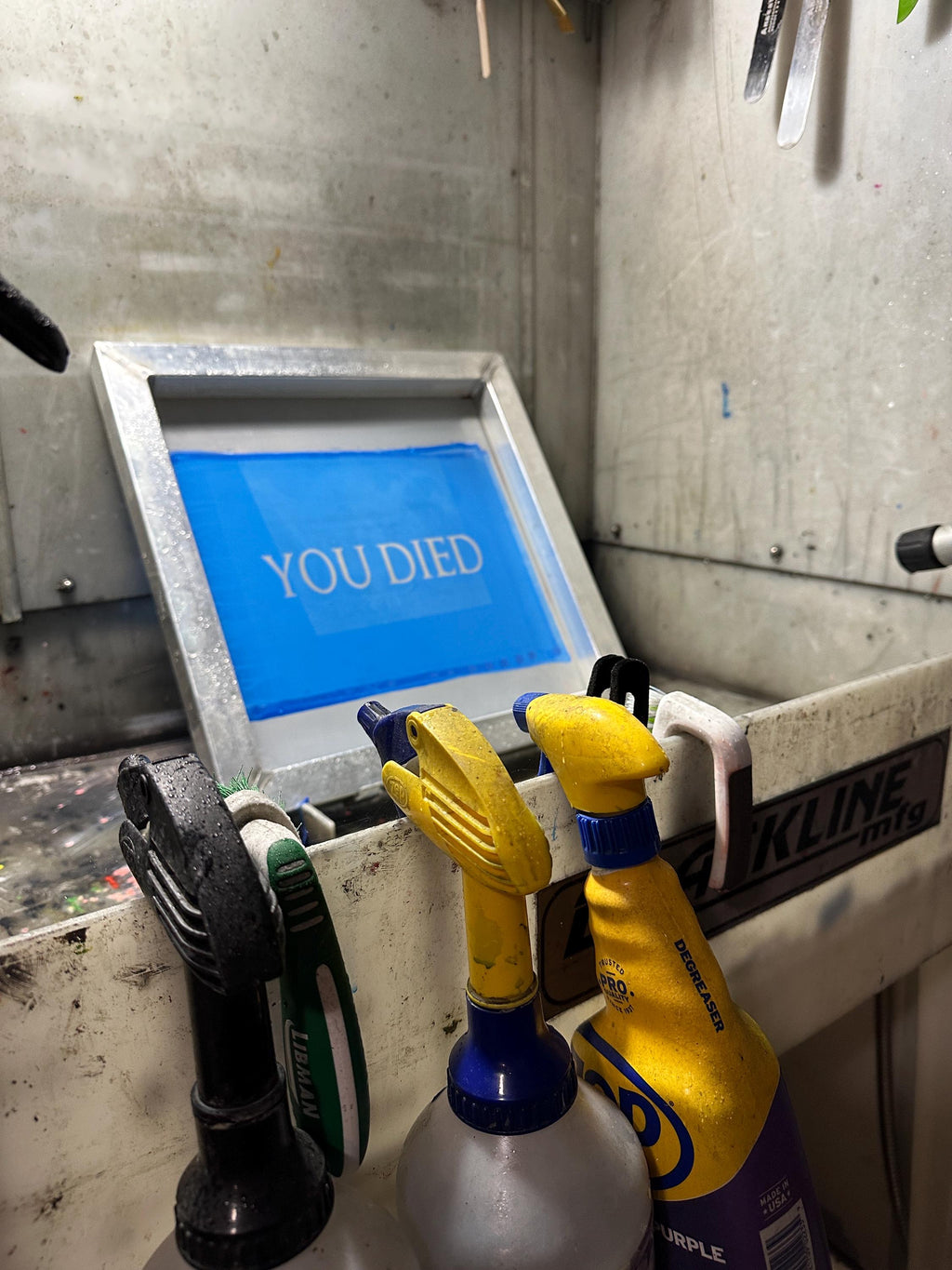 A silkscreen frame in a washout booth with a design that reads "YOU DIED" exposed in the blue emulsion