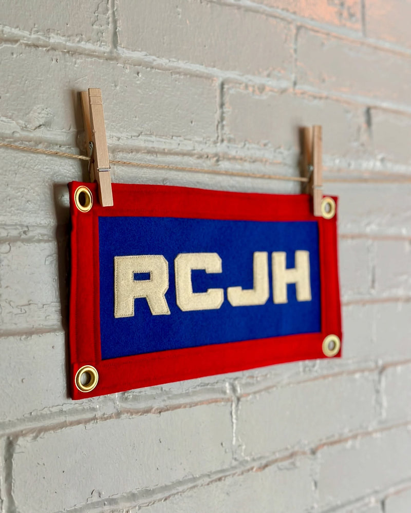 Felt banner with red border and 'RCJH' text on a blue background, hanging against a white brick wall.