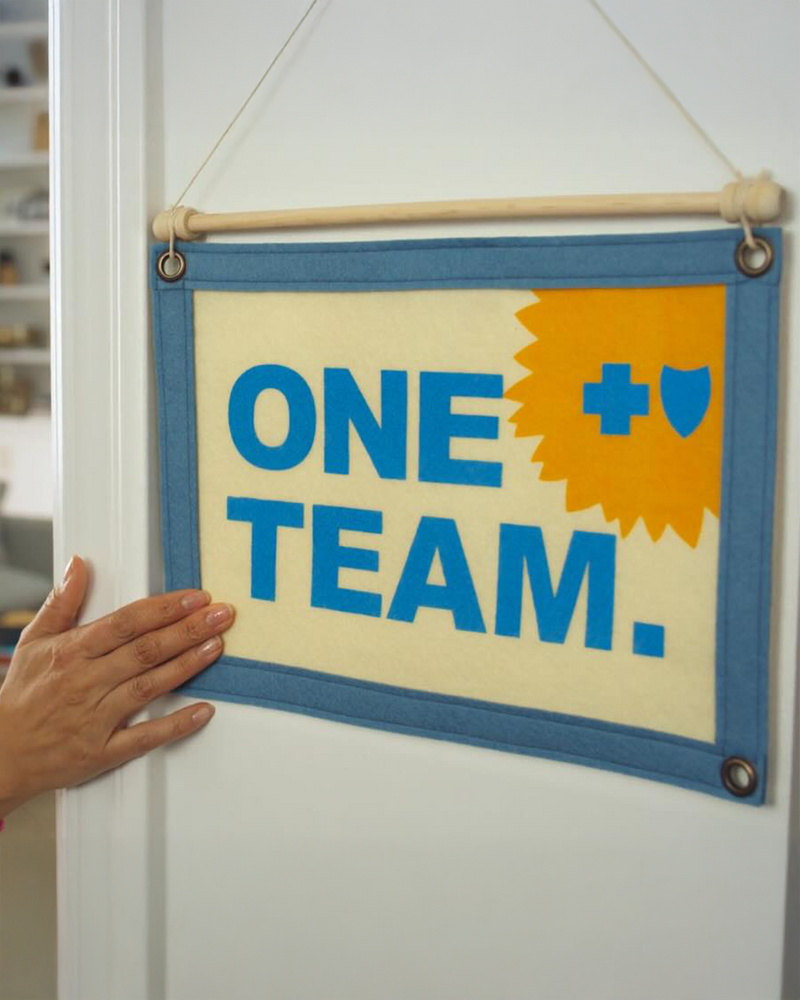 Hand touching a banner with 'ONE TEAM.' text on a white wall, as featured in a Blue Cross Blue Shield of Kansas commercial