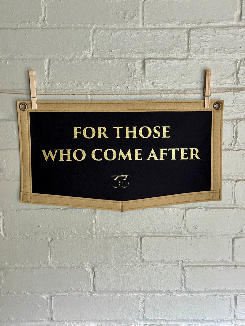 Black felt banner with light brown trim and gold lettering reads "For those who come after"