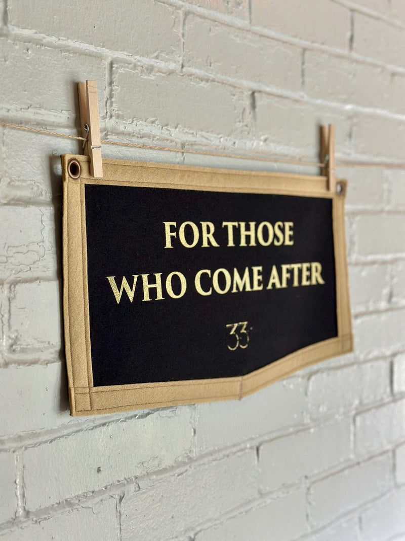Black felt banner with light brown trim and gold lettering reads "For those who come after"