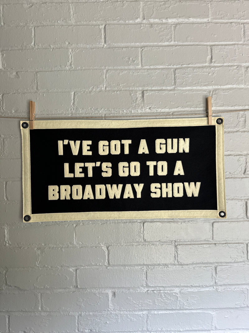 Black felt banner with text 'I've got a gun, let's go to a Broadway show' on a white brick wall.