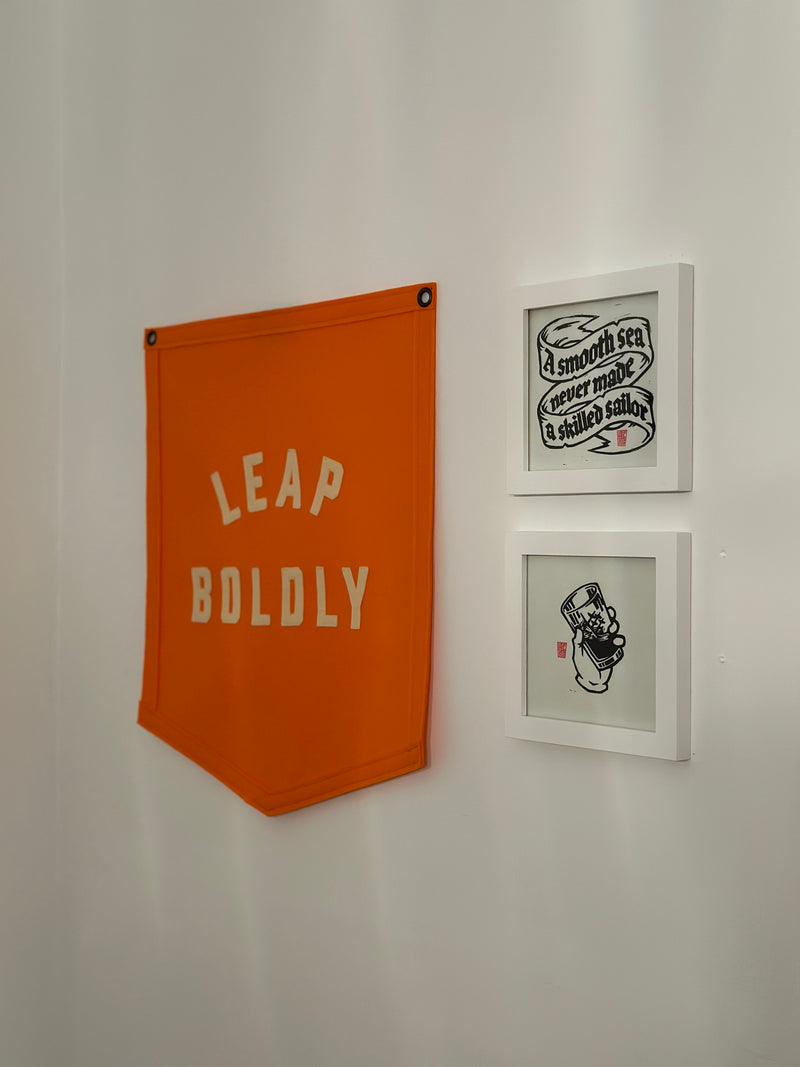 An orange banner reading LEAP BOLDLY hanging on a white wall alongside other framed artwork.