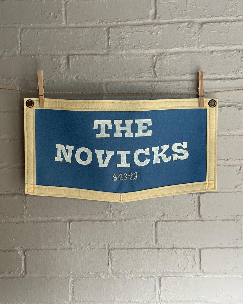 Blue sign with 'THE NOVICKS' and a chain stitched wedding date hung on a white brick wall.