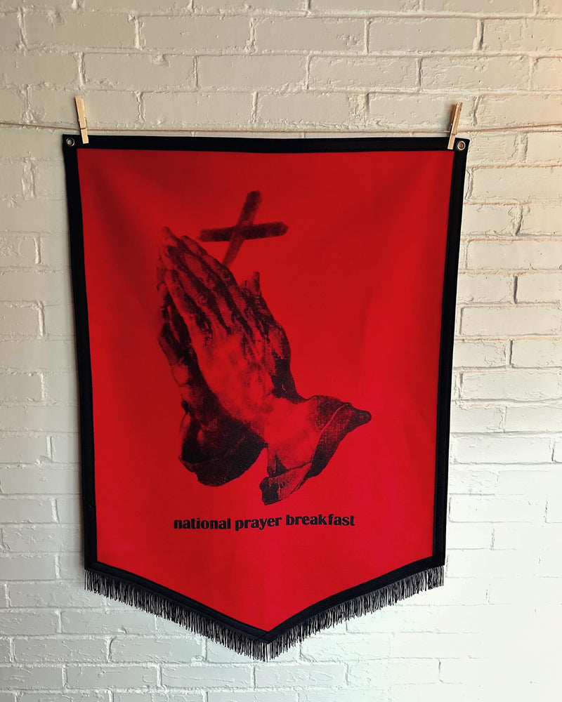 Red felt banner with black praying hands and cross design and the words 'National prayer breakfast' hanging on a brick wall.