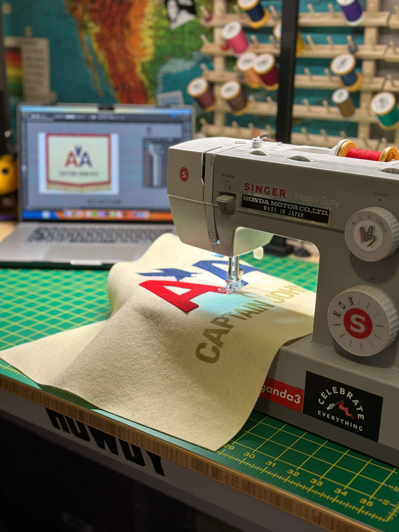 Sewing machine on a table with cutting mat and a banner in progress, with a laptop displaying the banner mockup in the background.