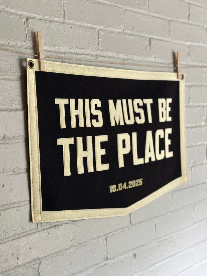 Pentagonal black felt banner with cream trim and cream sewn letters reading "This must be the place"