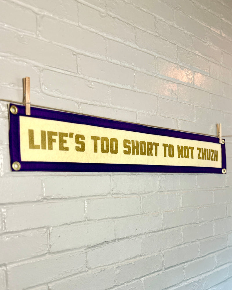 Decorative felt banner with text 'LIFE'S TOO SHORT TO NOT ZHUZZ' on a white brick wall.