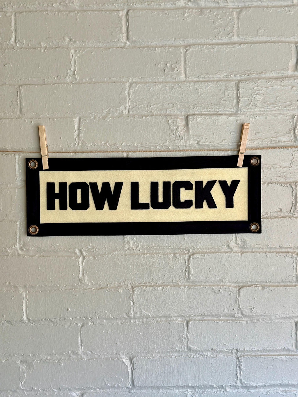 A cream felt banner with black stitched letters that read 'HOW LUCKY' hanging from two wooden clothespins against a white brick wall.