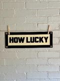 A cream felt banner with black stitched letters that read 'HOW LUCKY' hanging from two wooden clothespins against a white brick wall.
