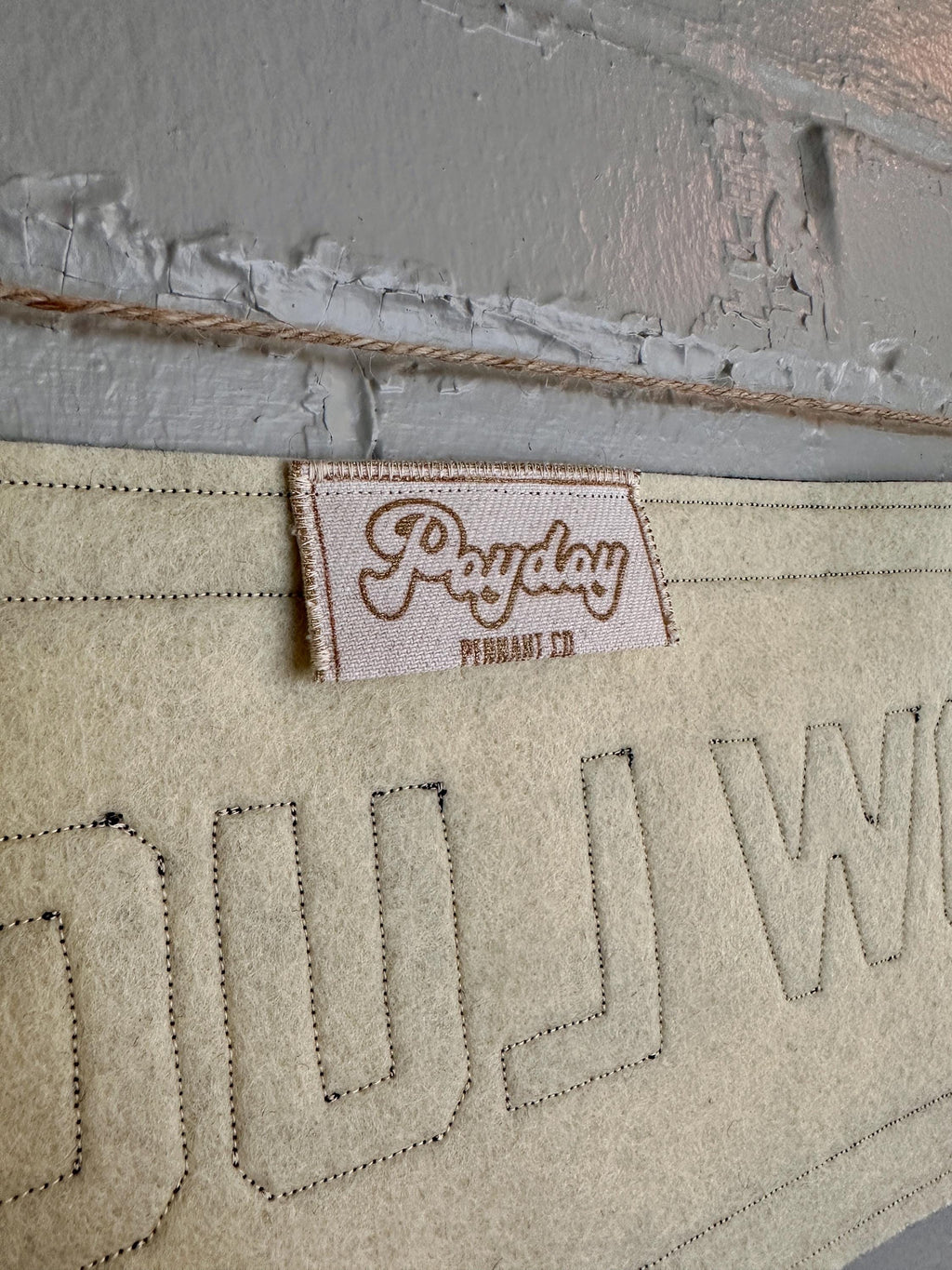 The back of a cream felt banner with black stitched letters that read 'HOW LUCKY' hanging from two wooden clothespins against a white brick wall.