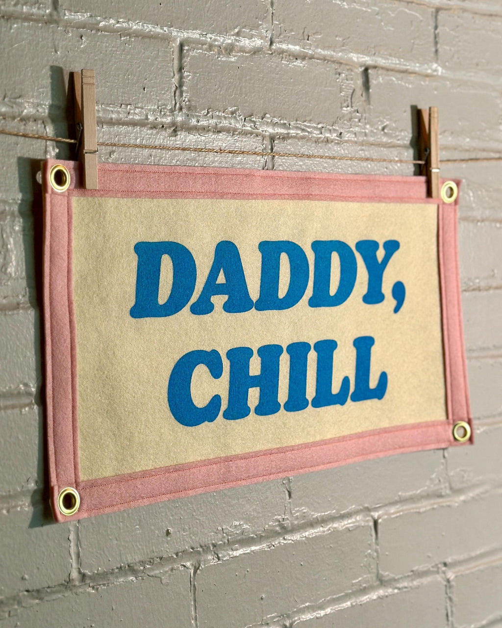 The side of a felt banner with cream felt background, pink felt trim and blue screenprinted text that reads 'DADDY, CHILL' hanging from a white brick wall with two wooden clothespins.