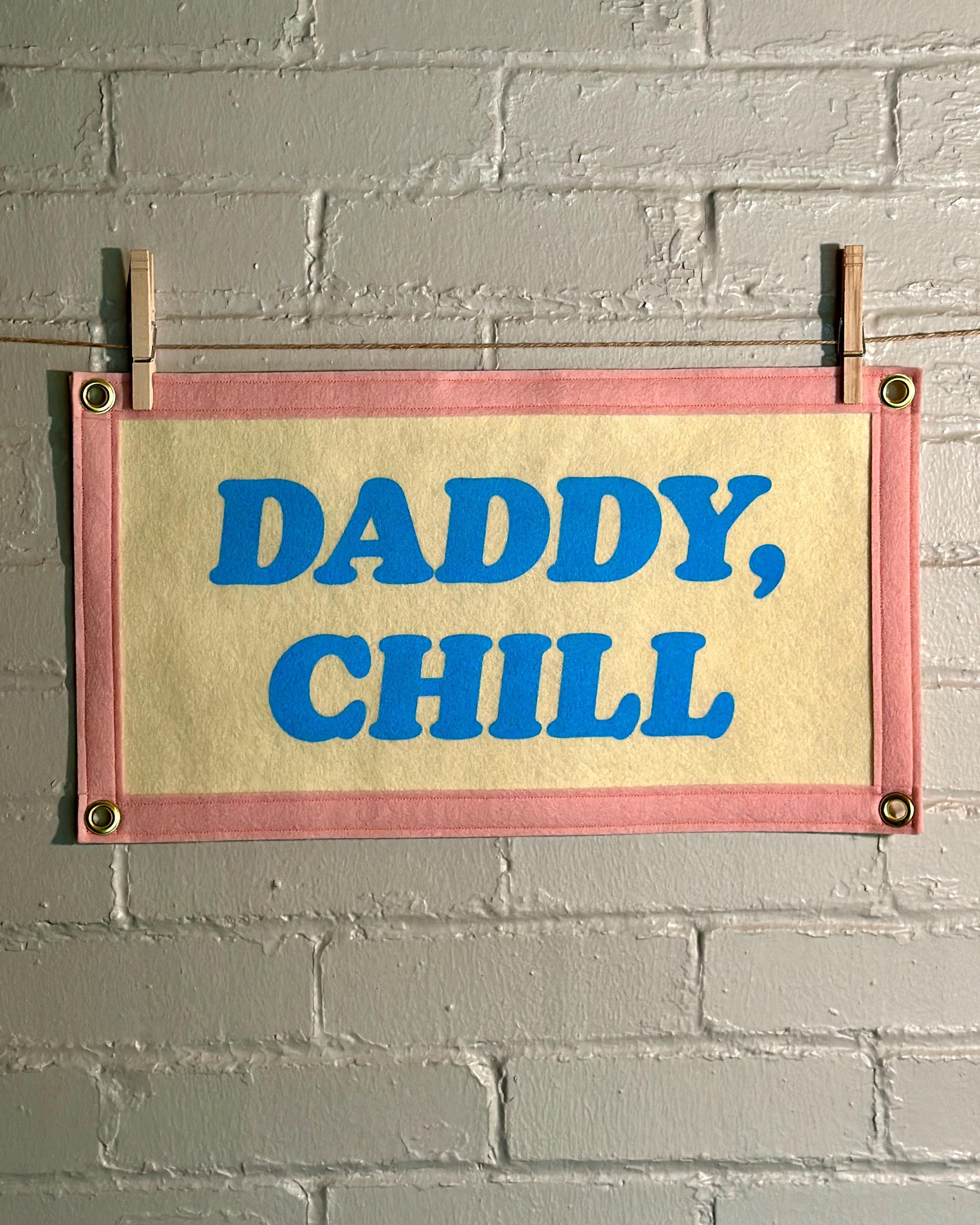 A felt banner with cream felt background, pink felt trim, and blue screenprinted text that reads 'DADDY, CHILL' hanging from a white brick wall with two wooden clothespins.