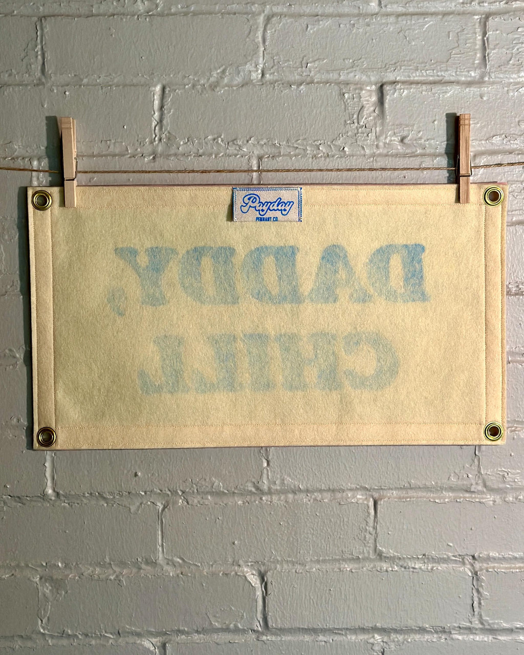 The back of a felt banner with cream felt background, pink felt trim and blue screenprinted text that reads 'DADDY, CHILL' hanging from a white brick wall with two wooden clothespins.