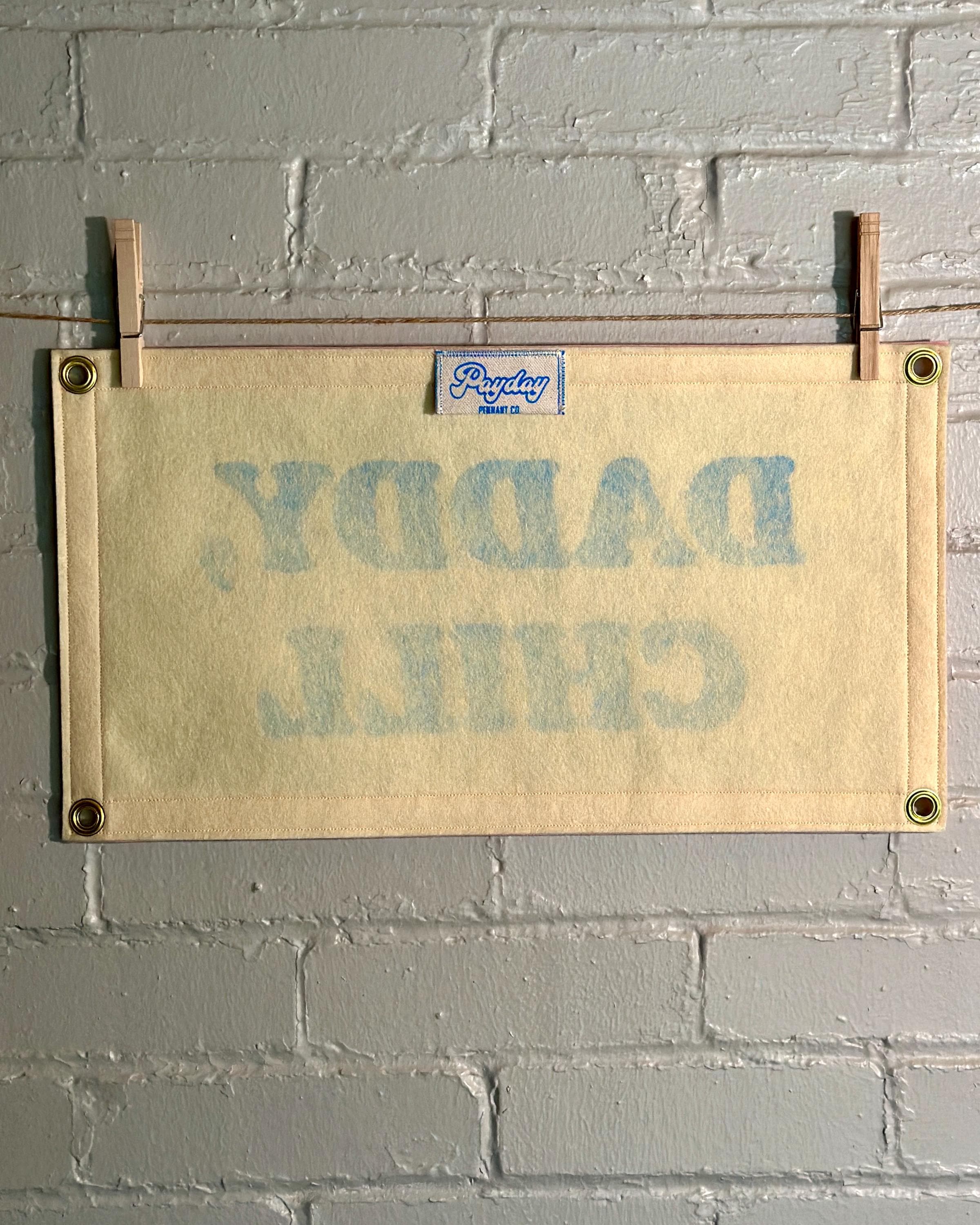 The back of a felt banner with cream felt background, pink felt trim and blue screenprinted text that reads 'DADDY, CHILL' hanging from a white brick wall with two wooden clothespins.