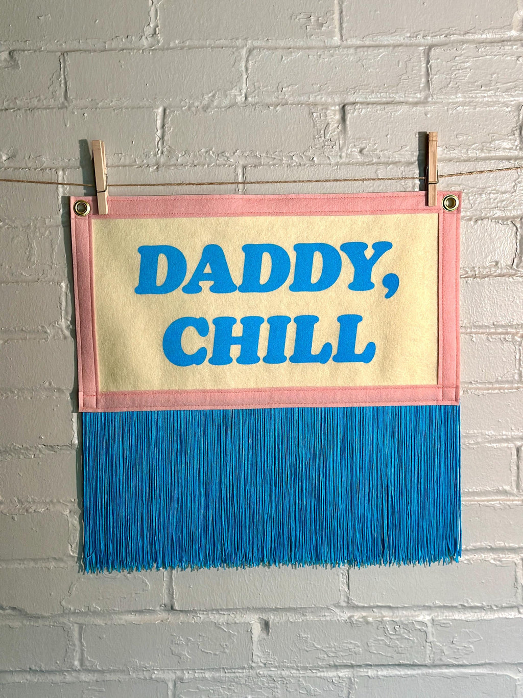 A felt banner with cream felt background, pink felt trim, long blue fringe at the bottom, and blue screenprinted text that reads 'DADDY, CHILL' hanging from a white brick wall with two wooden clothespins.