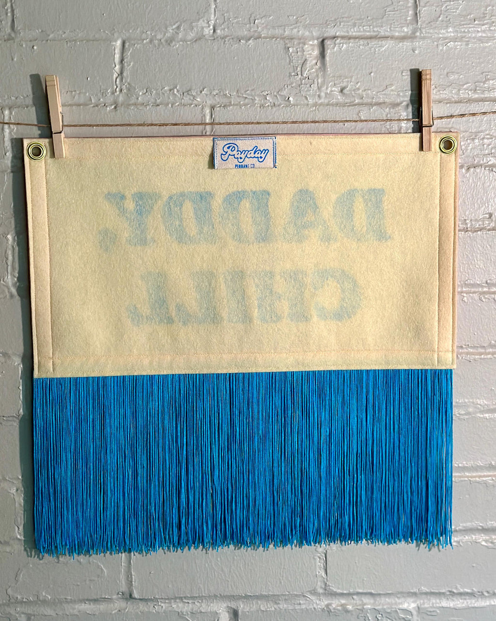 The back of a felt banner with cream felt background, pink felt trim, long blue fringe at the bottom, and blue screenprinted text that reads 'DADDY, CHILL' hanging from a white brick wall with two wooden clothespins.