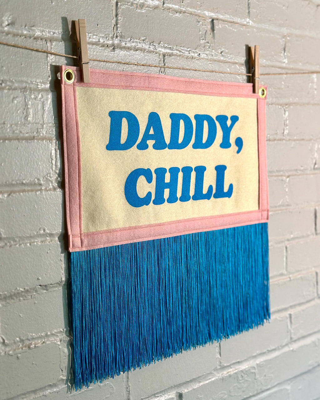 A felt banner with cream felt background, pink felt trim, long blue fringe at the bottom, and blue screenprinted text that reads 'DADDY, CHILL' hanging from a white brick wall with two wooden clothespins.