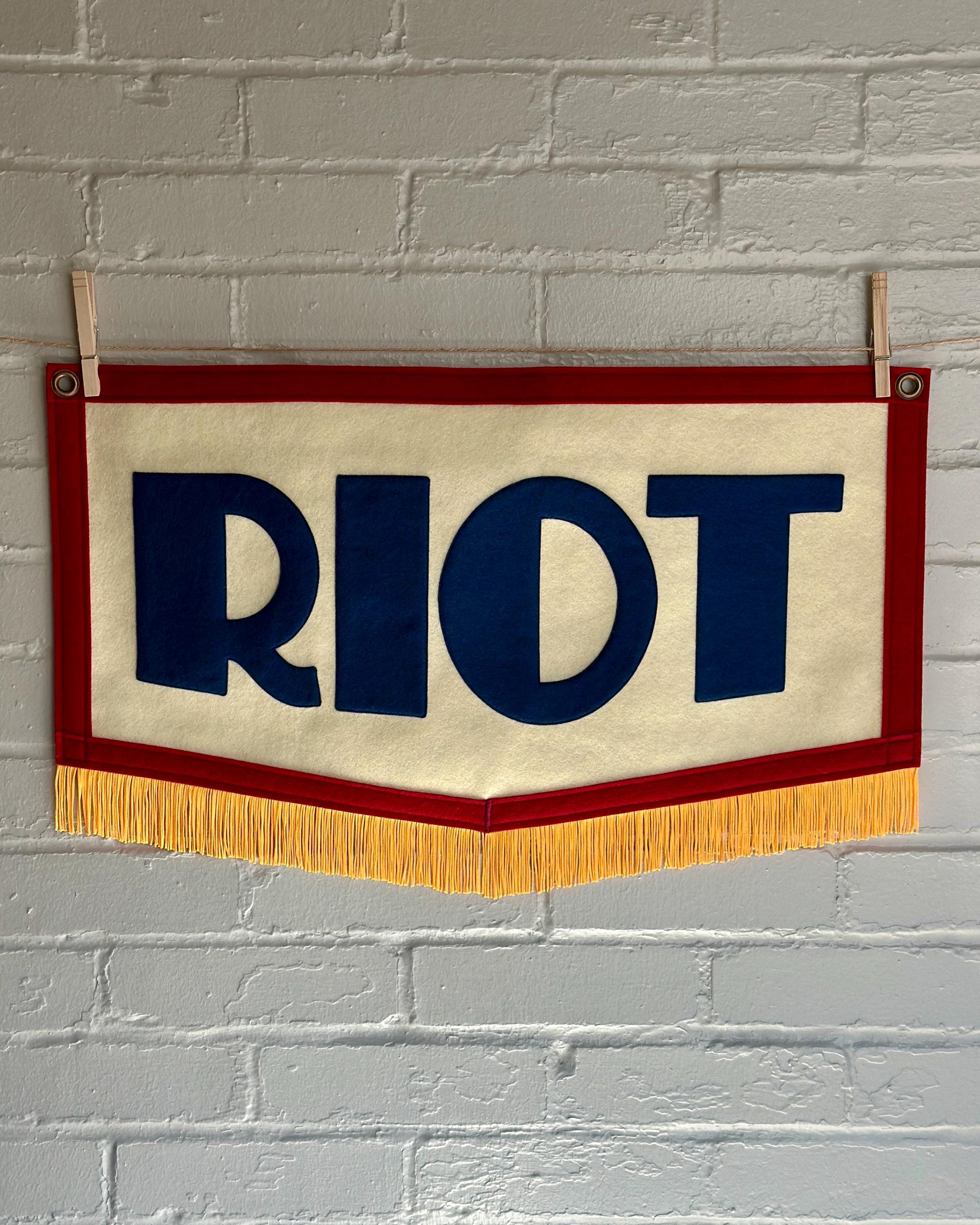 A red, white and blue felt banner with flag gold trim and appliqué letters that read RIOT