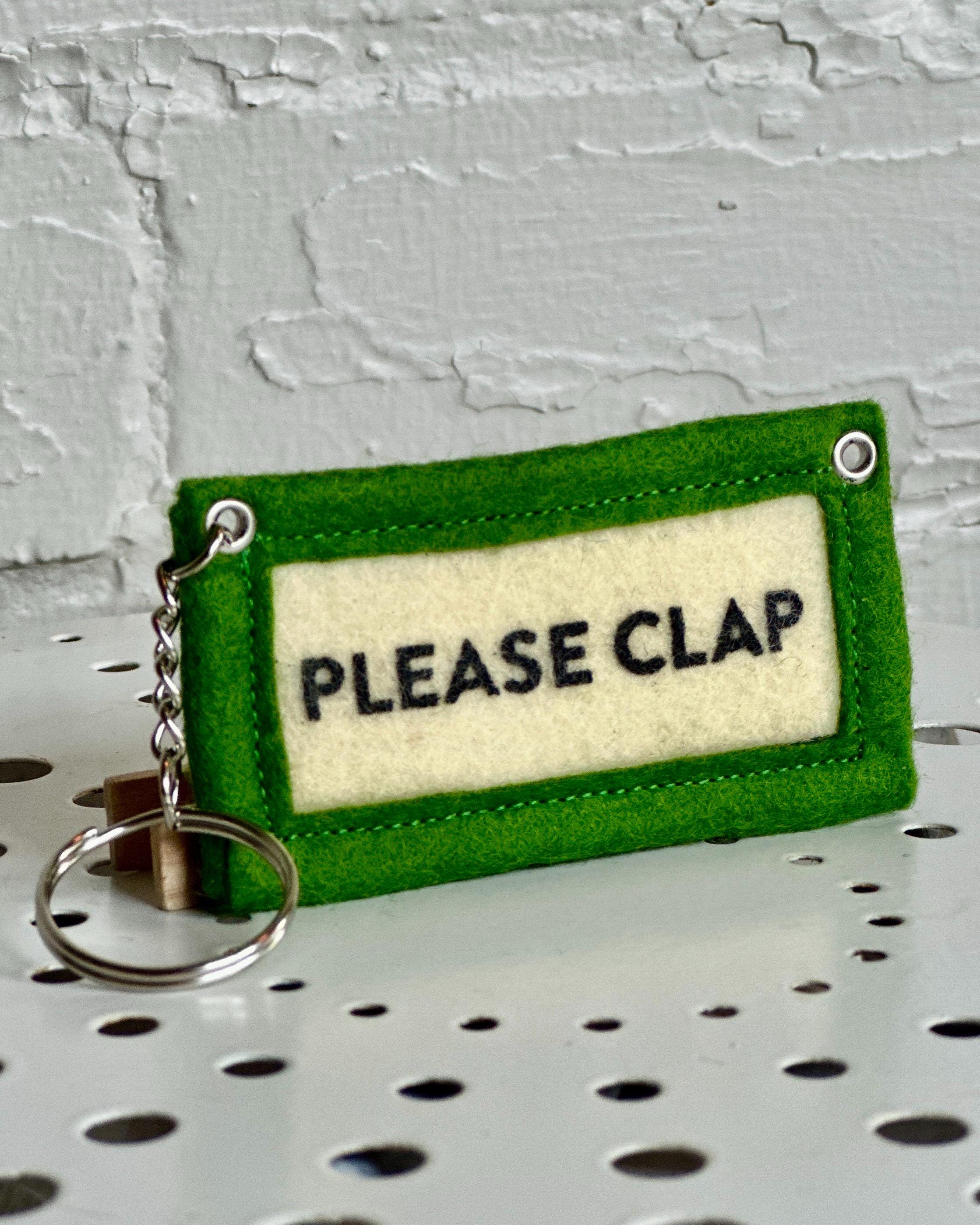 Green felt keychain with 'PLEASE CLAP' text in front of a white brick wall